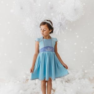 May include: A young girl wearing a light blue dress with a silver tiara. The dress features a light blue bodice with silver trim and a darker blue waist with a snowflake design. The girl is standing in a cloud-like setting.