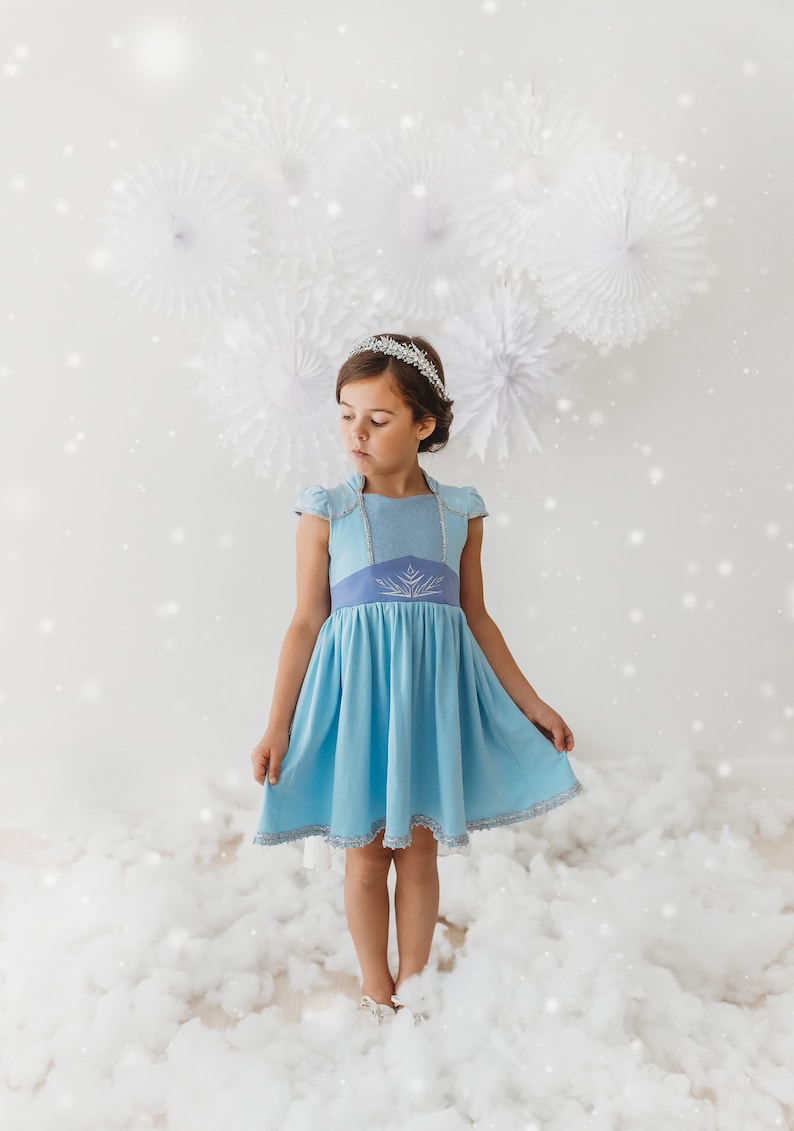 May include: A young girl wearing a light blue dress with a silver tiara. The dress features a light blue bodice with silver trim and a darker blue waist with a snowflake design. The girl is standing in a cloud-like setting.