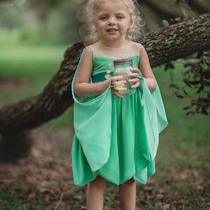 Pu&ograve; includere: Una bambina con un vestito da fata verde con maniche fluttuanti e corpetto beige. Tiene in mano un barattolo pieno di luci scintillanti. L'abito ha una gonna a strati ed &egrave; abbinato a sandali beige. Lo sfondo &egrave; una scena all'aperto sfocata.