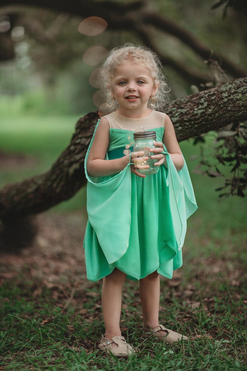 Pu&ograve; includere: Una bambina con un vestito da fata verde con maniche fluttuanti e corpetto beige. Tiene in mano un barattolo pieno di luci scintillanti. L'abito ha una gonna a strati ed &egrave; abbinato a sandali beige. Lo sfondo &egrave; una scena all'aperto sfocata.