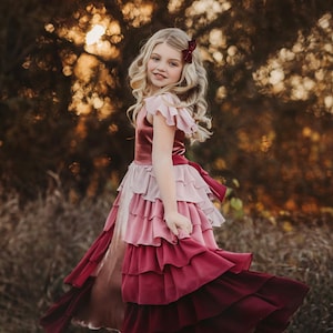 A girl is wearing the Yule Love all of the ruffles Maxi Dress in shades of pink and red. There are pink flutter sleeves. The bodice is a pink mauve and leads into cascading layers chiffon that start with dusty pink and flow down into a deep maroon.