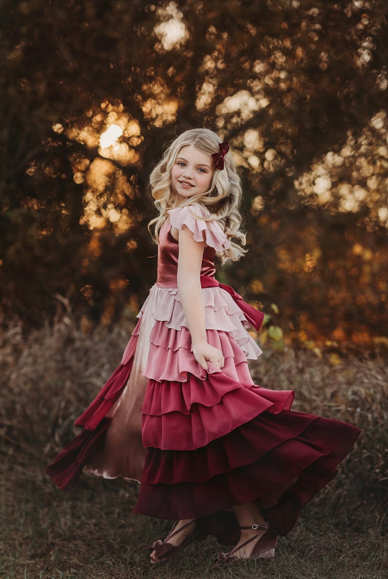 A girl is wearing the Yule Love all of the ruffles Maxi Dress in shades of pink and red. There are pink flutter sleeves. The bodice is a pink mauve and leads into cascading layers chiffon that start with dusty pink and flow down into a deep maroon.
