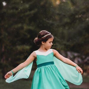 Pu&ograve; includere: Una giovane persona indossa un vestito turchese con dettagli bianchi e maniche fluttuanti. L'abito ha una cintura turchese scuro e una gonna svasata. Una fascia dorata e braccialetti turchesi completano l'outfit. Lo sfondo &egrave; verde sfocato.