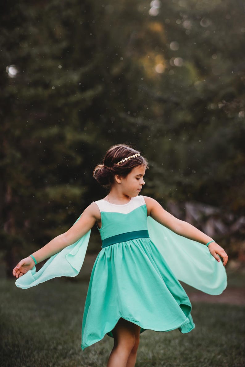 Pu&ograve; includere: Una giovane persona indossa un vestito turchese con dettagli bianchi e maniche fluttuanti. L'abito ha una cintura turchese scuro e una gonna svasata. Una fascia dorata e braccialetti turchesi completano l'outfit. Lo sfondo &egrave; verde sfocato.