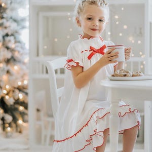 A young girl is wearing The Original Clara Nutcracker Christmas Gown Short Sleeve in White, featuring red accents, a bow, and a ruffled hem from the Christmas Lounge Collection. The buttery soft fabric is CPSC certified.
