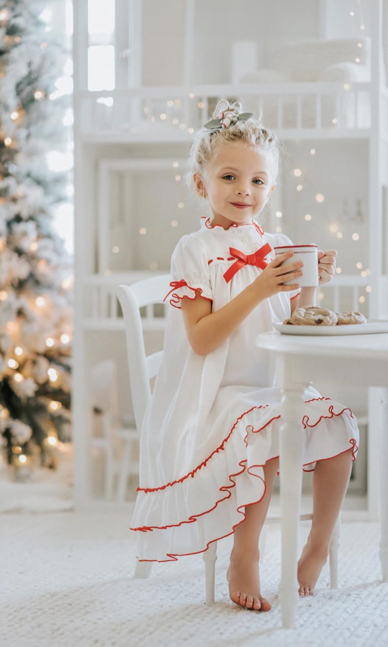 A young girl is wearing The Original Clara Nutcracker Christmas Gown Short Sleeve in White, featuring red accents, a bow, and a ruffled hem from the Christmas Lounge Collection. The buttery soft fabric is CPSC certified.