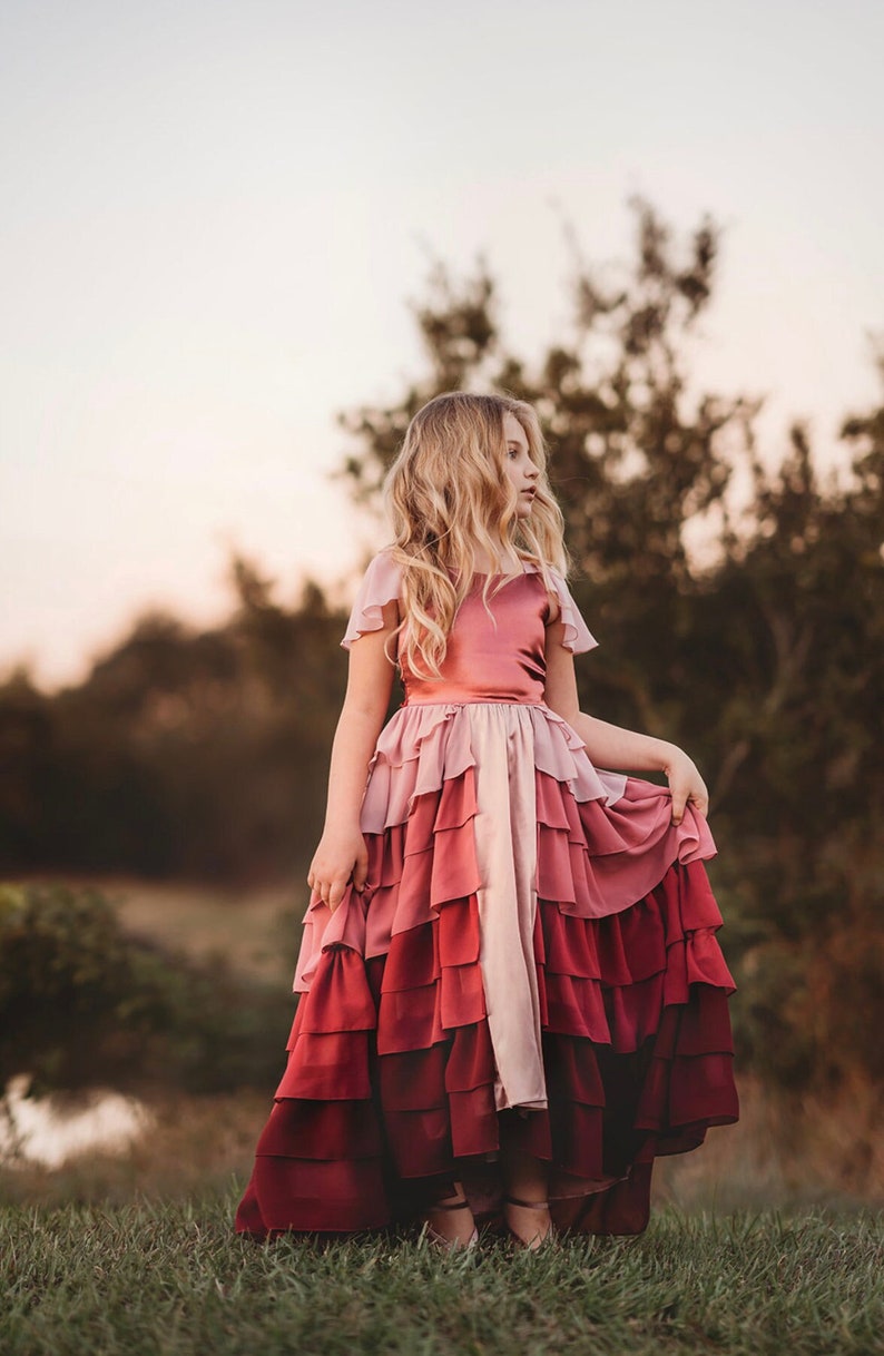 A girl is wearing the Yule Love all of the ruffles Maxi Dress in shades of pink and red. There are pink flutter sleeves. The bodice is a pink mauve and leads into cascading layers chiffon that start with dusty pink and flow down into a deep maroon.
