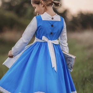 A young girl stands wearing the Red Rose Princess Village Dress from the Fairy Godmother Collection, reminiscent of Disney's Belle from "Beauty and the Beast." The white sleeves are long. The dress has a bight blue chest and skirt.