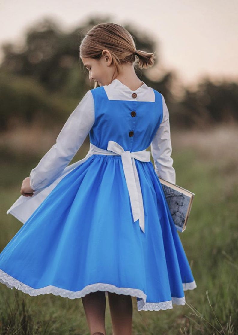 A young girl stands wearing the Red Rose Princess Village Dress from the Fairy Godmother Collection, reminiscent of Disney's Belle from "Beauty and the Beast." The white sleeves are long. The dress has a bight blue chest and skirt.