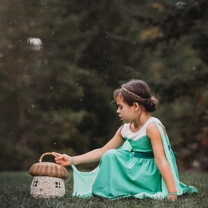 Pu&ograve; includere: Una giovane persona con un vestito verde acqua e una fascia dorata &egrave; seduta sull'erba accanto a un cestino a forma di fungo. Il cestino ha una parte superiore intrecciata e una piccola base a forma di casa. Lo sfondo &egrave; verde sfocato.