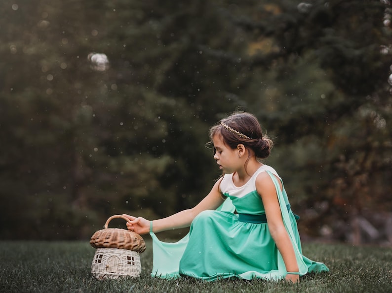 Pu&ograve; includere: Una giovane persona con un vestito verde acqua e una fascia dorata &egrave; seduta sull'erba accanto a un cestino a forma di fungo. Il cestino ha una parte superiore intrecciata e una piccola base a forma di casa. Lo sfondo &egrave; verde sfocato.