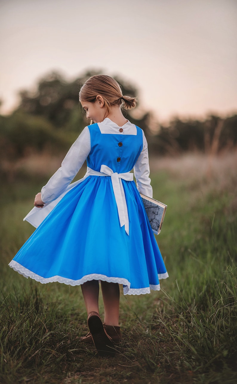 A young girl stands wearing the Red Rose Princess Village Dress from the Fairy Godmother Collection, reminiscent of Disney's Belle from "Beauty and the Beast." The white sleeves are long. The dress has a bight blue chest and skirt.