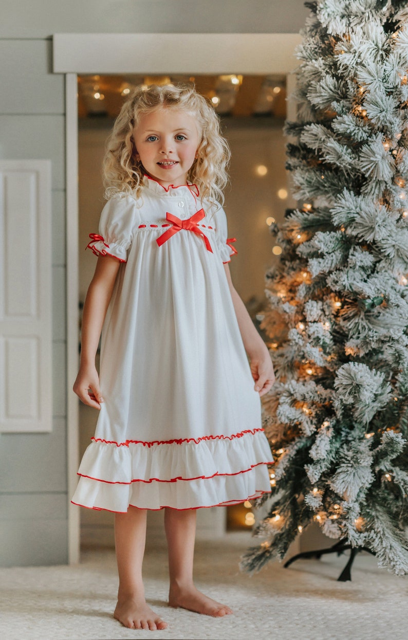 A young girl is wearing The Original Clara Nutcracker Christmas Gown Short Sleeve in White, featuring red accents, a bow, and a ruffled hem from the Christmas Lounge Collection. The buttery soft fabric is CPSC certified.