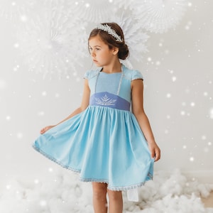May include: A light blue dress with a silver tiara. The dress features a light blue bodice with cap sleeves and a darker blue waistline with a silver snowflake design. The skirt is a lighter blue with silver trim.