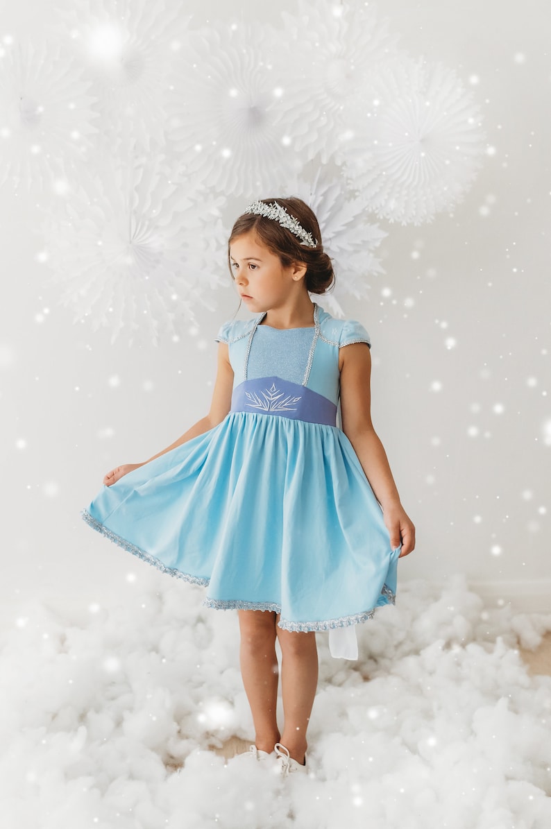 May include: A light blue dress with a silver tiara. The dress features a light blue bodice with cap sleeves and a darker blue waistline with a silver snowflake design. The skirt is a lighter blue with silver trim.