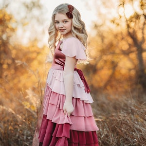 A girl is wearing the Yule Love all of the ruffles Maxi Dress in shades of pink and red. There are pink flutter sleeves. The bodice is a pink mauve and leads into cascading layers chiffon that start with dusty pink and flow down into a deep maroon.
