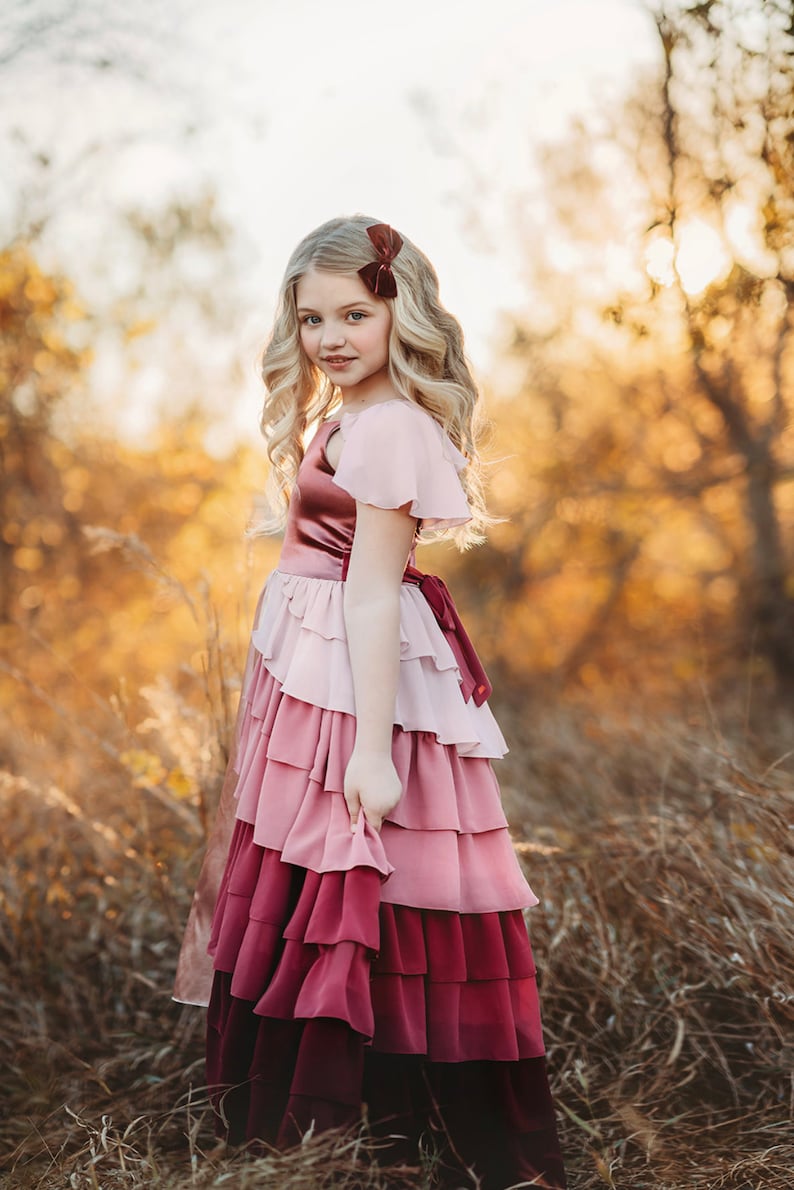 A girl is wearing the Yule Love all of the ruffles Maxi Dress in shades of pink and red. There are pink flutter sleeves. The bodice is a pink mauve and leads into cascading layers chiffon that start with dusty pink and flow down into a deep maroon.
