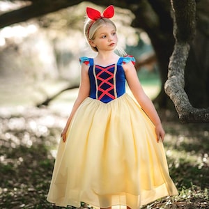 Könnte beinhalten: Ein bodenlanges Kleid mit einem blauen Samtoberteil mit roter Schnürung, kurzen Puffärmeln mit blauen und roten Akzenten und einem weiten gelben Rock. Das Outfit umfasst ein rotes Stirnband mit Schleife sowie dazu passende rote flache Schuhe.