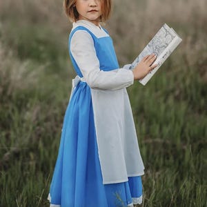 A young girl stands wearing the Red Rose Princess Village Dress from the Fairy Godmother Collection, reminiscent of Disney's Belle from "Beauty and the Beast." The white sleeves are long. The dress has a bight blue chest and skirt.