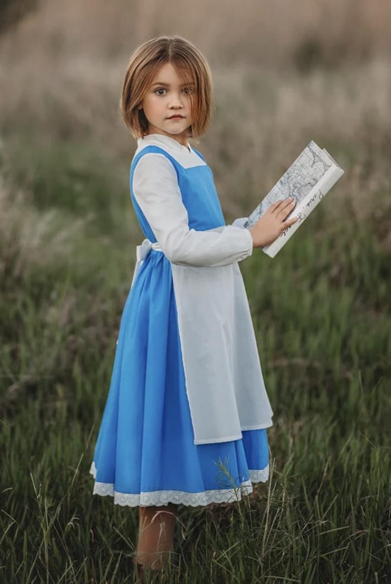 A young girl stands wearing the Red Rose Princess Village Dress from the Fairy Godmother Collection, reminiscent of Disney's Belle from "Beauty and the Beast." The white sleeves are long. The dress has a bight blue chest and skirt.
