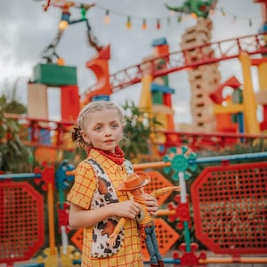 May include: A young person dressed in a yellow plaid shirt and vest, resembling a character from the movie "Toy Story", holds a Woody doll. The outfit includes a red bandana. The background features a colorful amusement park setting with a roller coaster.