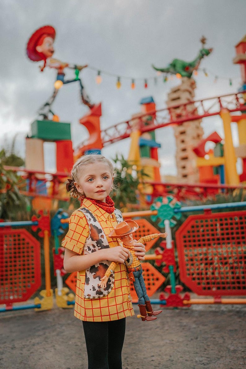 May include: A young person dressed in a yellow plaid shirt and vest, resembling a character from the movie "Toy Story", holds a Woody doll. The outfit includes a red bandana. The background features a colorful amusement park setting with a roller coaster.