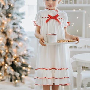 A young girl is wearing The Original Clara Nutcracker Christmas Gown Short Sleeve in White, featuring red accents, a bow, and a ruffled hem from the Christmas Lounge Collection. The buttery soft fabric is CPSC certified.