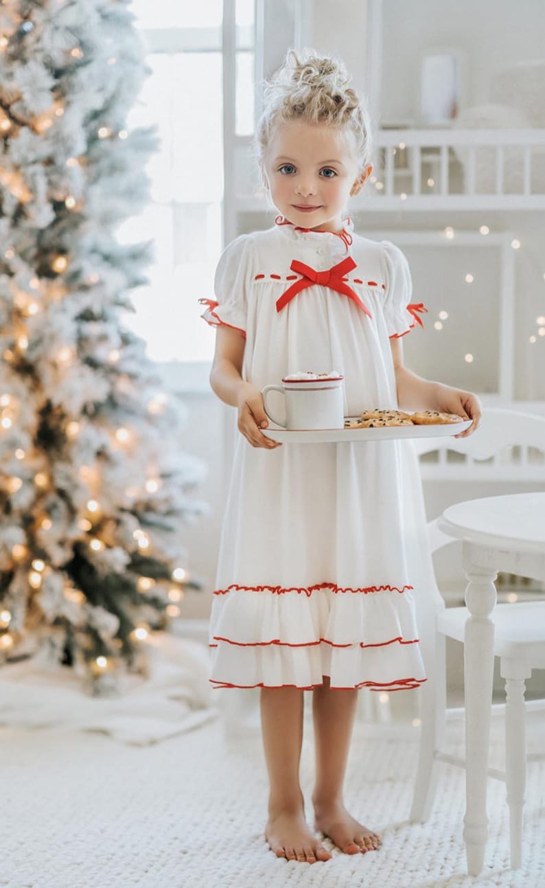 A young girl is wearing The Original Clara Nutcracker Christmas Gown Short Sleeve in White, featuring red accents, a bow, and a ruffled hem from the Christmas Lounge Collection. The buttery soft fabric is CPSC certified.