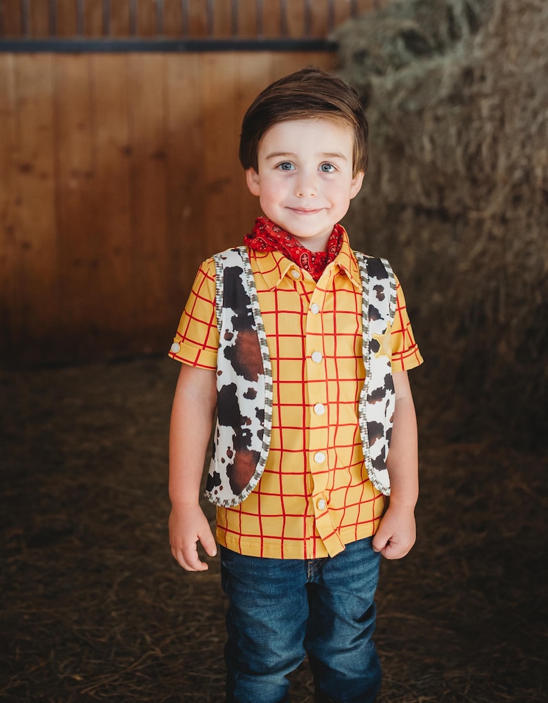 May include: A child wearing a cowboy costume, featuring a yellow plaid shirt with red lines, a cow-print vest, a red bandana, and blue jeans. The costume is reminiscent of a classic Western character.
