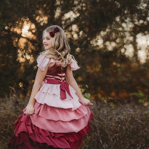 A girl is wearing the Yule Love all of the ruffles Maxi Dress in shades of pink and red. There are pink flutter sleeves. The bodice is a pink mauve and leads into cascading layers chiffon that start with dusty pink and flow down into a deep maroon.
