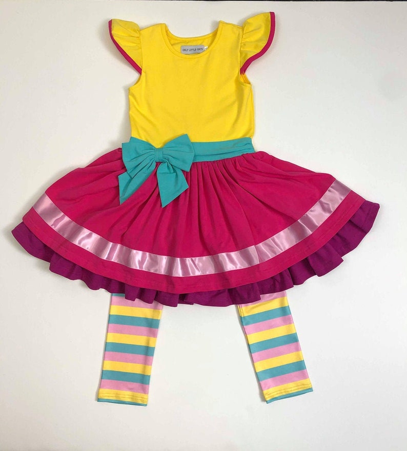 Pu&ograve; includere: Un vestito giallo senza maniche con un fiocco turchese e una gonna rosa con un nastro di raso rosa. L'abito &egrave; abbinato a leggings a righe in giallo, rosa e turchese.