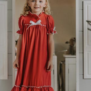 A young girl is wearing The Original Clara Nutcracker Christmas Gown Short Sleeve in RED, featuring White accents, a bow, and a ruffled hem from the Christmas Lounge Collection. The buttery soft fabric is CPSC certified.
