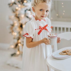 A young girl is wearing The Original Clara Nutcracker Christmas Gown Short Sleeve in White, featuring red accents, a bow, and a ruffled hem from the Christmas Lounge Collection. The buttery soft fabric is CPSC certified.