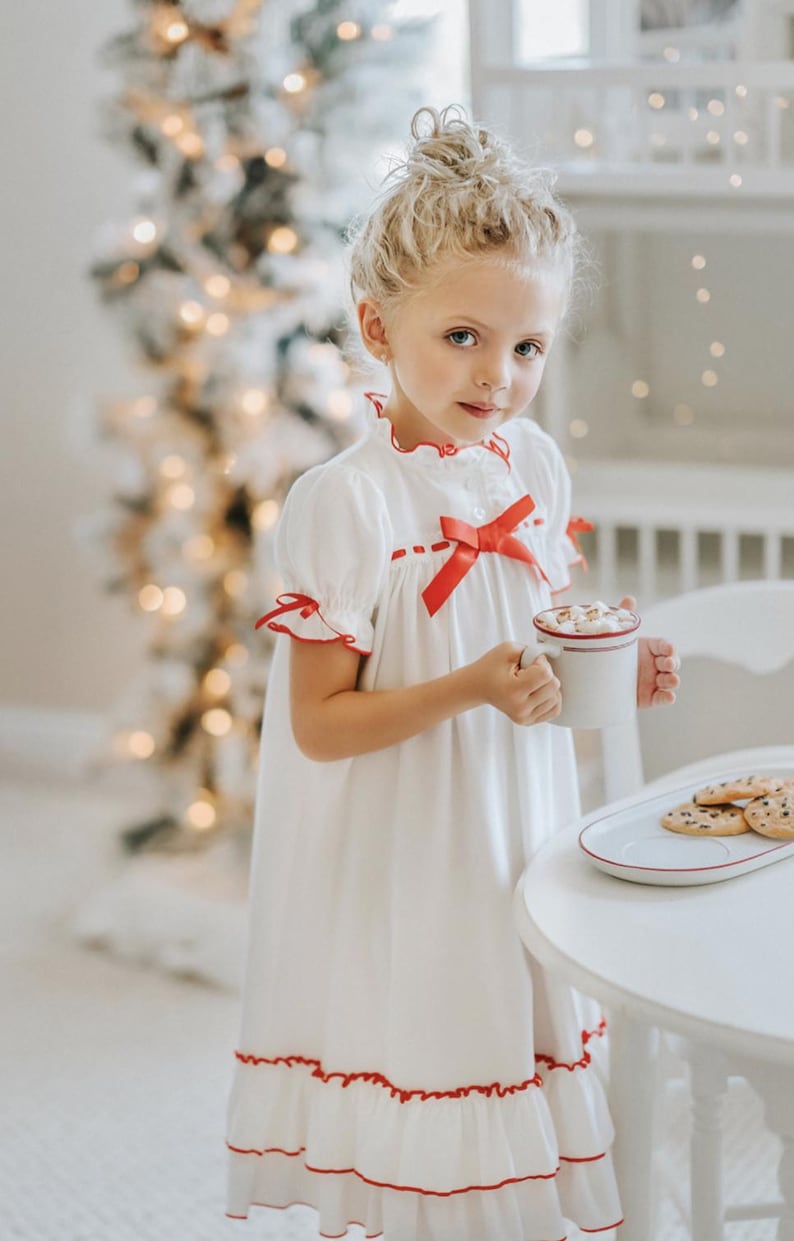 A young girl is wearing The Original Clara Nutcracker Christmas Gown Short Sleeve in White, featuring red accents, a bow, and a ruffled hem from the Christmas Lounge Collection. The buttery soft fabric is CPSC certified.