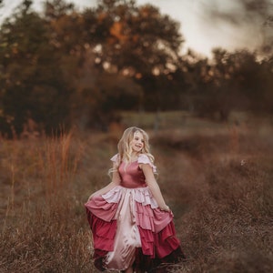 A girl is wearing the Yule Love all of the ruffles Maxi Dress in shades of pink and red. There are pink flutter sleeves. The bodice is a pink mauve and leads into cascading layers chiffon that start with dusty pink and flow down into a deep maroon.