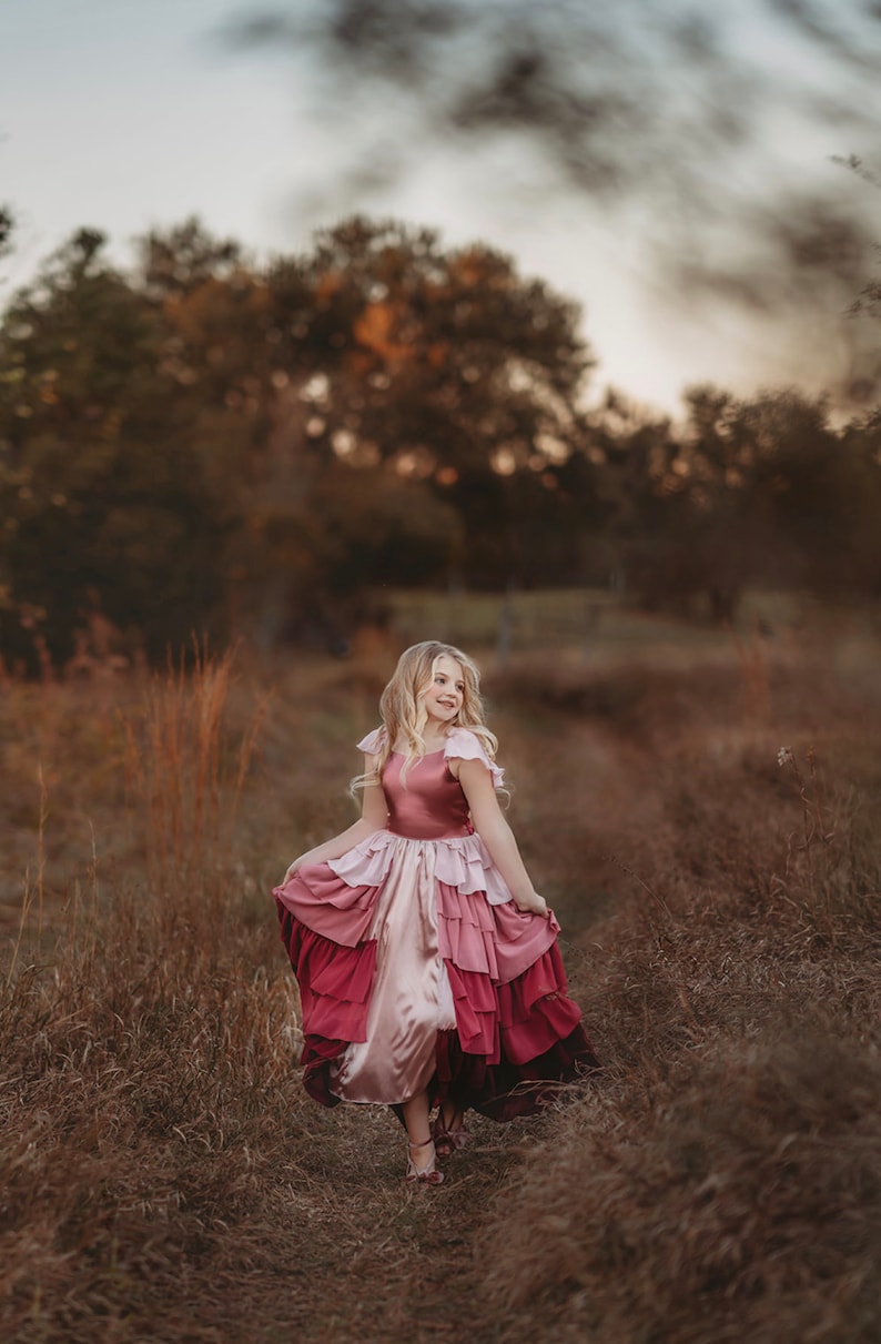 A girl is wearing the Yule Love all of the ruffles Maxi Dress in shades of pink and red. There are pink flutter sleeves. The bodice is a pink mauve and leads into cascading layers chiffon that start with dusty pink and flow down into a deep maroon.