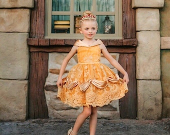 May include: A golden yellow dress featuring an off-the-shoulder neckline, a lace-overlay bodice, and a skirt with draped fabric accents. The dress is paired with a metallic tiara adorned with small red gems, and gold-colored strappy sandals.