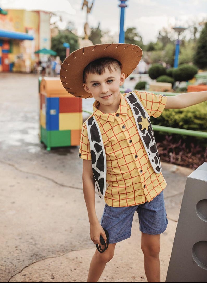 Puede incluir: Un ni&ntilde;o peque&ntilde;o vestido como Woody de Toy Story. Lleva una camisa de cuadros amarillos y rojos con un sombrero de vaquero marr&oacute;n, un chaleco blanco y negro y pantalones cortos de mezclilla azul.
