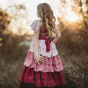 A girl is wearing the Yule Love all of the ruffles Maxi Dress in shades of pink and red. There are pink flutter sleeves. The bodice is a pink mauve and leads into cascading layers chiffon that start with dusty pink and flow down into a deep maroon.