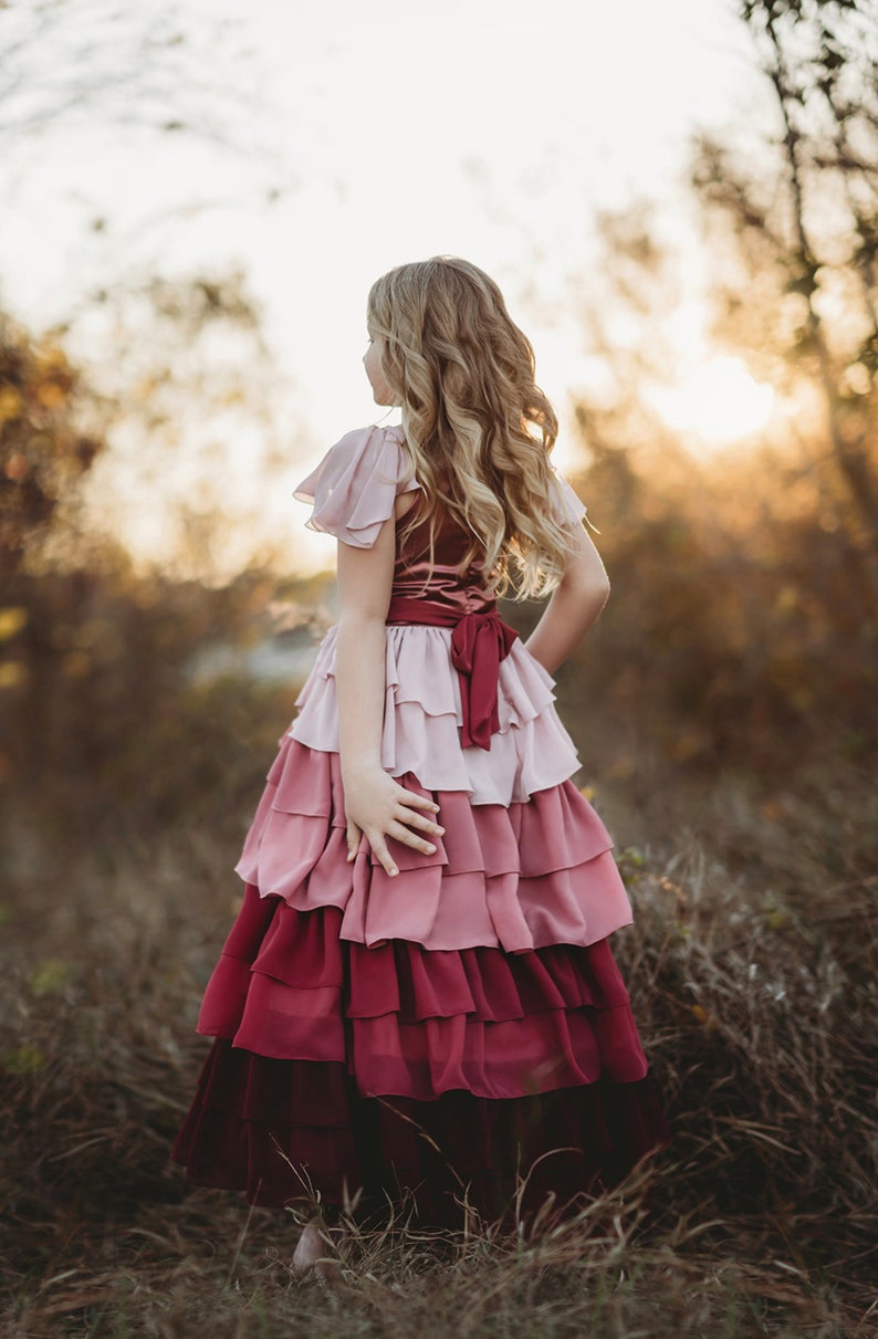 A girl is wearing the Yule Love all of the ruffles Maxi Dress in shades of pink and red. There are pink flutter sleeves. The bodice is a pink mauve and leads into cascading layers chiffon that start with dusty pink and flow down into a deep maroon.