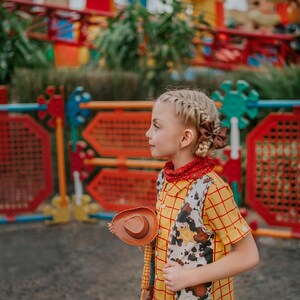 May include: A young person dressed in a Woody costume, featuring a yellow plaid shirt, a cow-print vest, and a brown cowboy hat. The outfit includes a red bandana and black pants. The background shows a colorful amusement park.