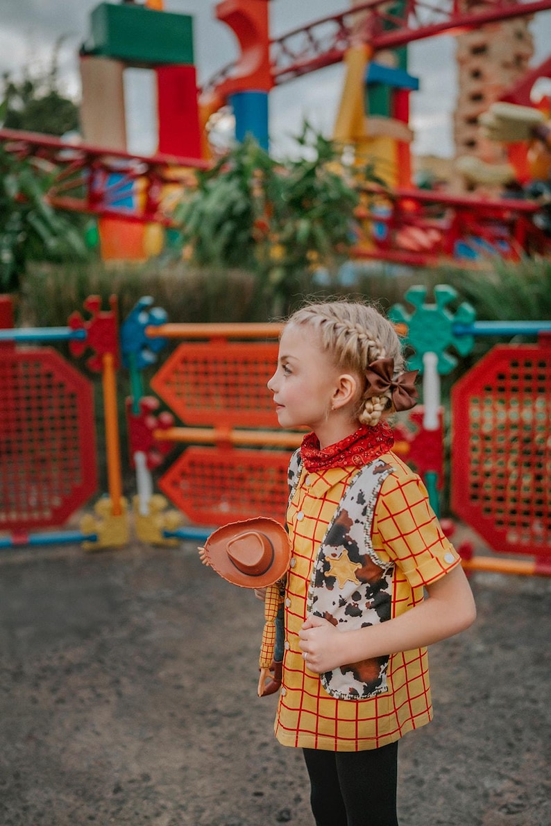 May include: A young person dressed in a Woody costume, featuring a yellow plaid shirt, a cow-print vest, and a brown cowboy hat. The outfit includes a red bandana and black pants. The background shows a colorful amusement park.
