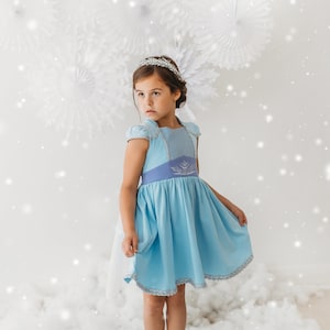 May include: A young girl wearing a light blue dress with a silver tiara. The dress has a light blue bodice with a darker blue yoke and a silver trim. The skirt is a lighter blue with a silver trim. The background has white paper decorations and a snowy effect.
