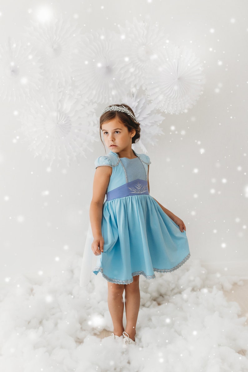 May include: A young girl wearing a light blue dress with a silver tiara. The dress has a light blue bodice with a darker blue yoke and a silver trim. The skirt is a lighter blue with a silver trim. The background has white paper decorations and a snowy effect.