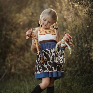 Puede incluir: Una ni&ntilde;a peque&ntilde;a con un vestido azul y blanco con una falda de estampado de vaca sostiene dos mu&ntilde;ecas de juguete vestidas como Woody y Jessie de la franquicia de pel&iacute;culas Toy Story.