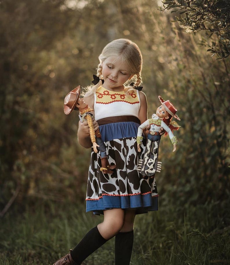 Puede incluir: Una ni&ntilde;a peque&ntilde;a con un vestido azul y blanco con una falda de estampado de vaca sostiene dos mu&ntilde;ecas de juguete vestidas como Woody y Jessie de la franquicia de pel&iacute;culas Toy Story.