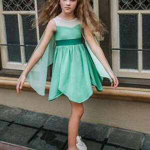Pu&ograve; includere: Una ragazza indossa un vestito verde menta con maniche a mantella trasparenti. L'abito ha una cintura verde scuro e uno scollo bianco. Indossa scarpe bianche con dettagli traforati. Lo sfondo presenta una finestra di legno.