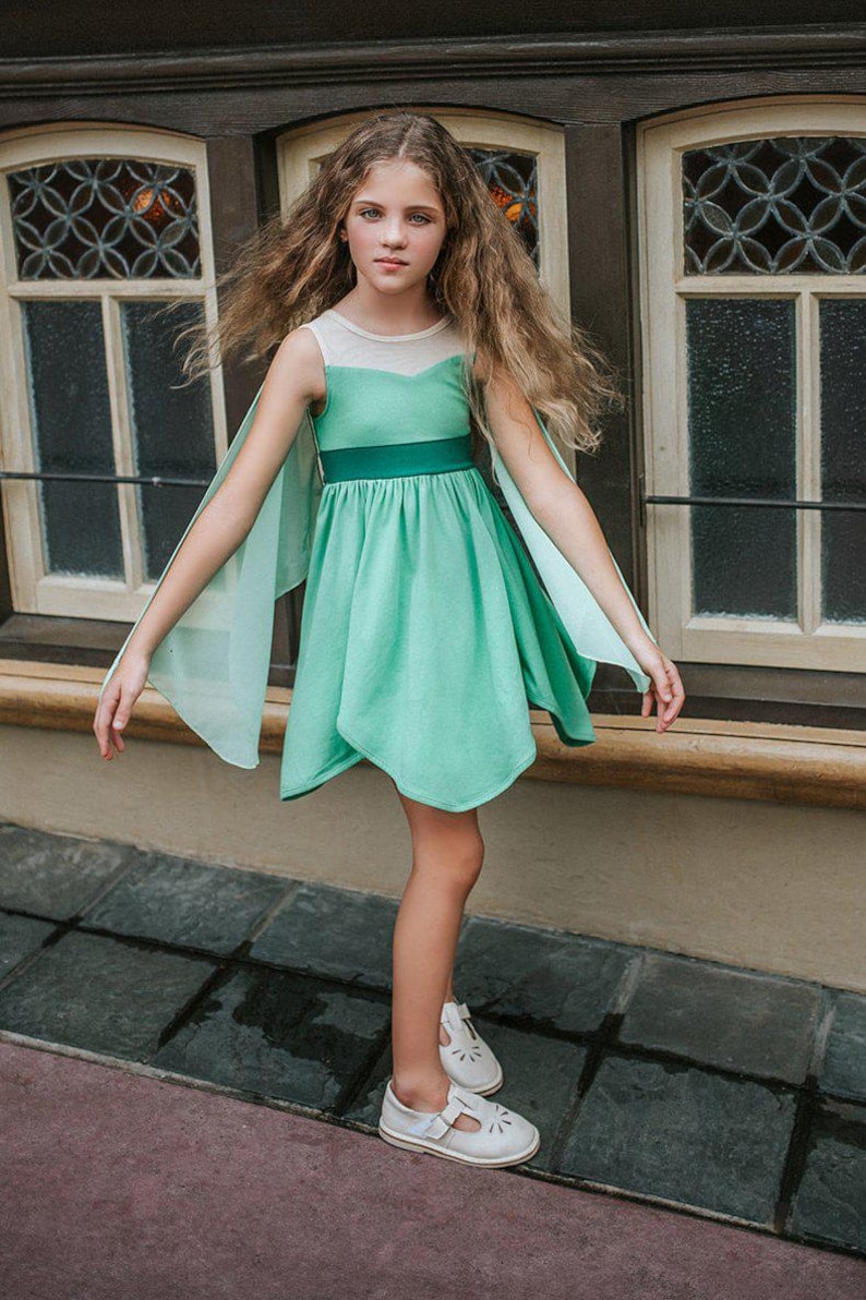 Pu&ograve; includere: Una ragazza indossa un vestito verde menta con maniche a mantella trasparenti. L'abito ha una cintura verde scuro e uno scollo bianco. Indossa scarpe bianche con dettagli traforati. Lo sfondo presenta una finestra di legno.