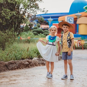 Puede incluir: Dos ni&ntilde;os disfrazados de Buzz Lightyear y Woody de Toy Story. La ni&ntilde;a lleva un vestido blanco con un disfraz de Buzz Lightyear encima. El ni&ntilde;o lleva una camisa de cuadros amarilla y roja con un chaleco marr&oacute;n y un sombrero de vaquero. Est&aacute;n de pie en un camino frente a un edificio colorido.