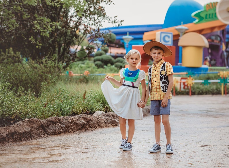 Puede incluir: Dos ni&ntilde;os disfrazados de Buzz Lightyear y Woody de Toy Story. La ni&ntilde;a lleva un vestido blanco con un disfraz de Buzz Lightyear encima. El ni&ntilde;o lleva una camisa de cuadros amarilla y roja con un chaleco marr&oacute;n y un sombrero de vaquero. Est&aacute;n de pie en un camino frente a un edificio colorido.